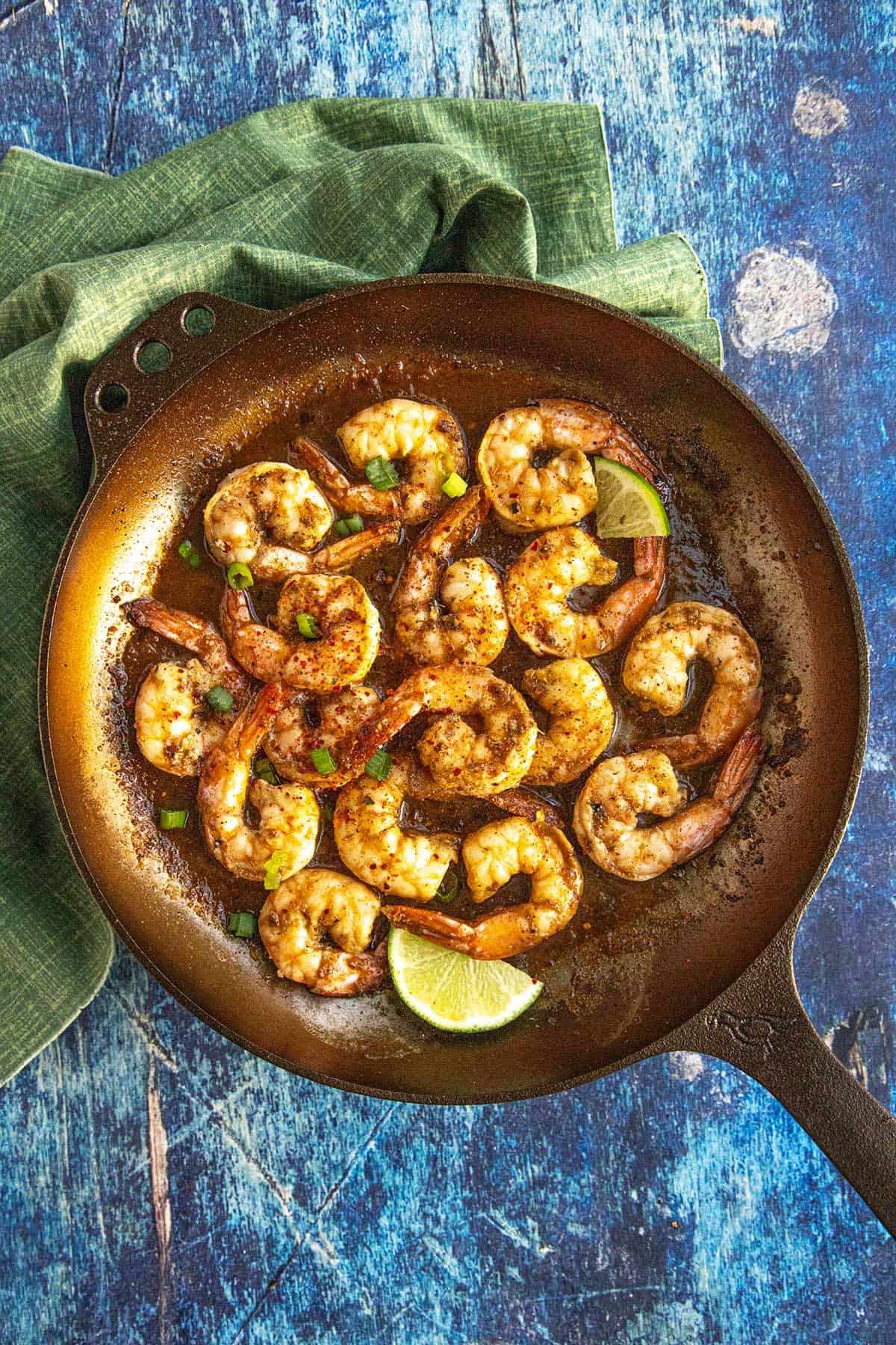 A brown skillet containing cooked, seasoned jerk shrimp garnished with green herbs and lime wedges sits on a green cloth atop a blue textured surface.