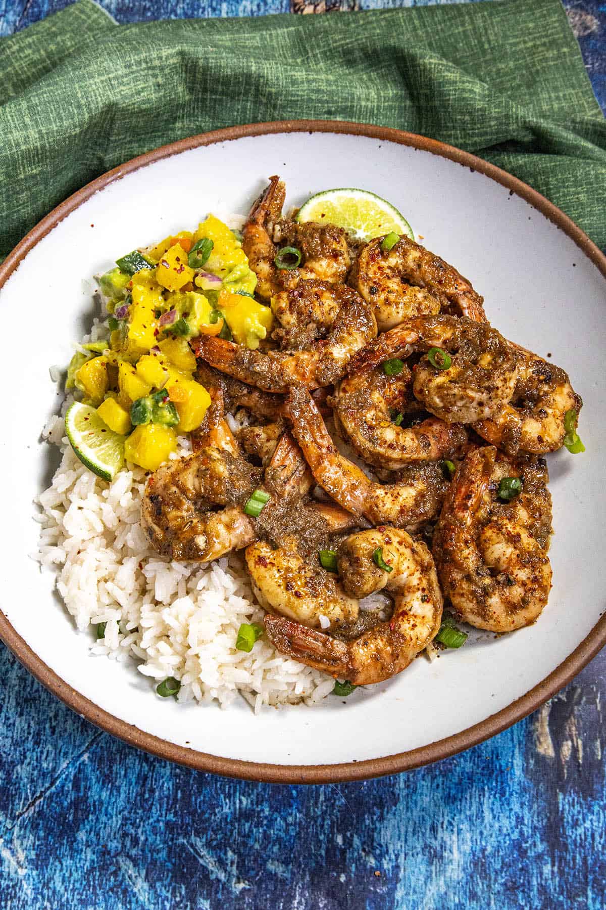 A bowl of jerk shrimp with coconut rice, topped with vibrant mango salsa, garnished with lime wedges and chopped green onions, sits on a blue surface beside a green cloth napkin.