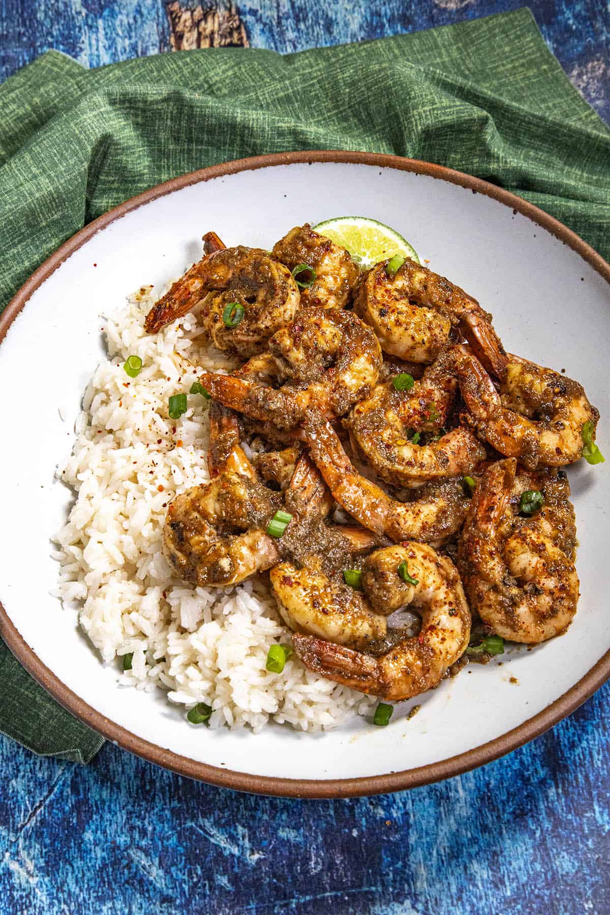 A bowl of white coconut rice topped with flavorful jerk shrimp, garnished with chopped green onions and a lime wedge, set on a blue textured surface with a green cloth napkin.