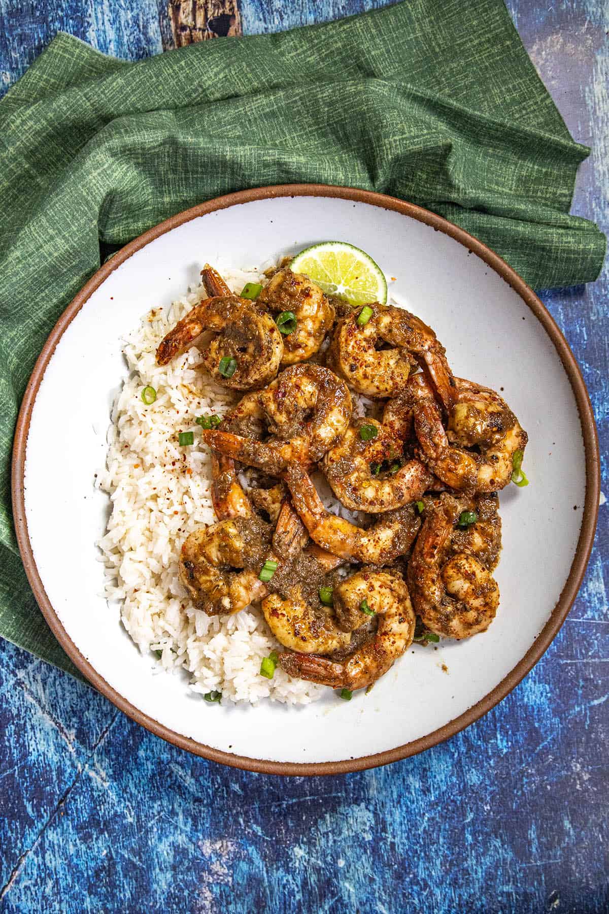 A plate of jerk shrimp served with coconut rice and garnished with green onions and a lime wedge, set on a blue textured surface with a green napkin.