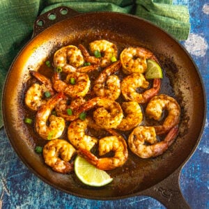 A cast iron skillet filled with Spicy Jerk Shrimp, garnished with chopped green onions and lime wedges, rests on a textured blue surface with a green cloth nearby.