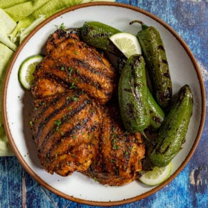 A plate with grilled, seasoned chicken breasts marinated in a zesty Mexican Chicken Marinade, charred jalapeño peppers, and lime slices, garnished with herbs, on a blue rustic surface.