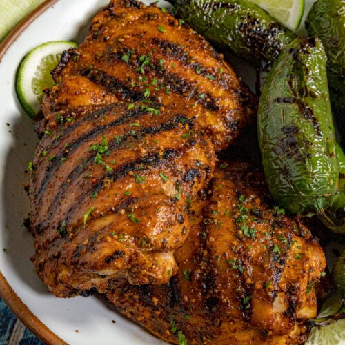 Grilled chicken thighs, marinated in a zesty Mexican Chicken Marinade, display char marks and are garnished with herbs, served alongside grilled jalape&ntilde;o peppers and lime slices.