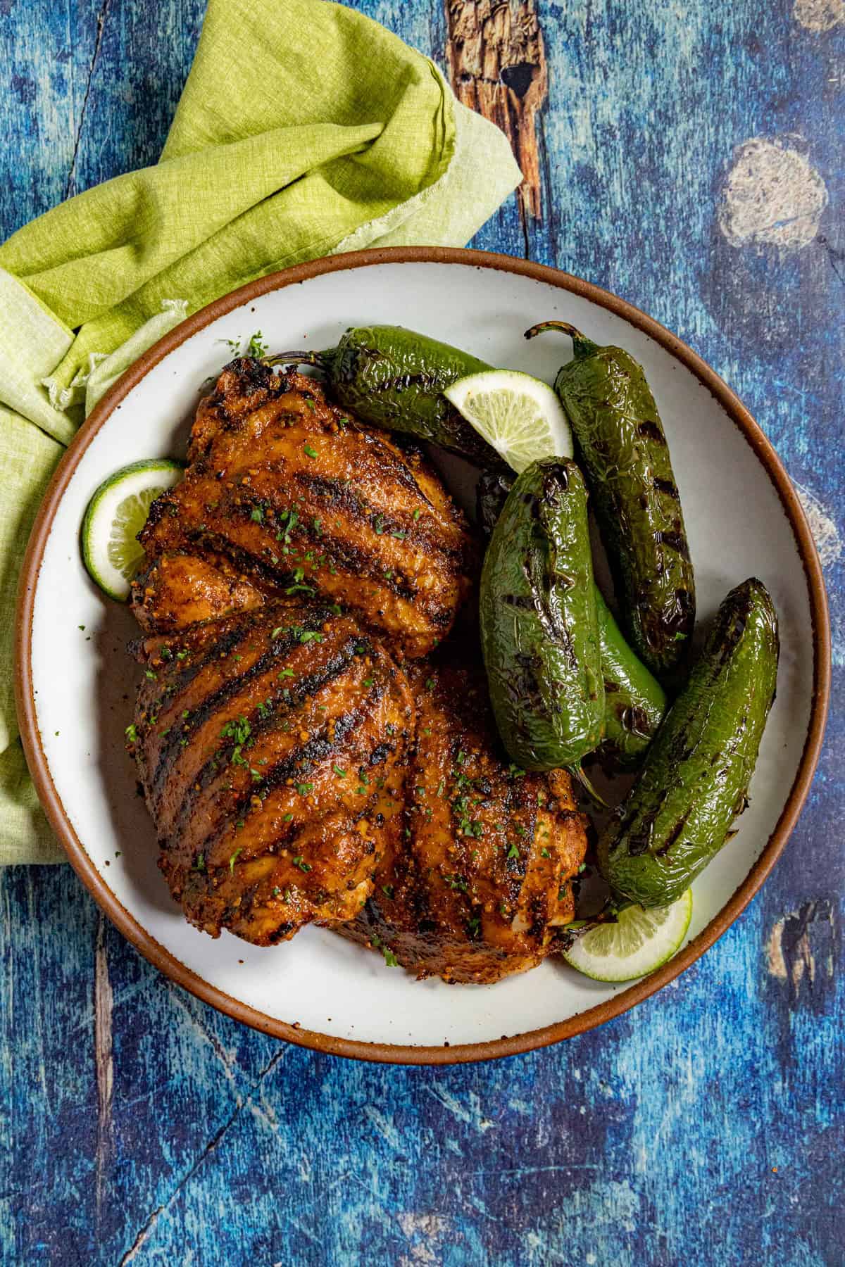 A plate with grilled, Mexican Chicken Marinade-seasoned chicken thighs and roasted jalapeños, garnished with lime slices, on a blue rustic surface. A folded green napkin sits beside the plate.
