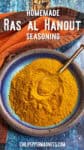 A blue bowl filled with yellow Ras el Hanout seasoning powder and a small brass spoon sits on a rustic blue surface with an orange and black textured cloth. Text above reads, Homemade Ras Al Hanout Recipe.