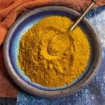 A blue-rimmed ceramic bowl filled with vibrant yellow Homemade Ras el Hanout, with a golden spoon resting on top, sits on a blue and orange textured surface.