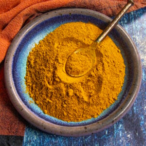 A blue-rimmed ceramic bowl filled with vibrant yellow Homemade Ras el Hanout, with a golden spoon resting on top, sits on a blue and orange textured surface.