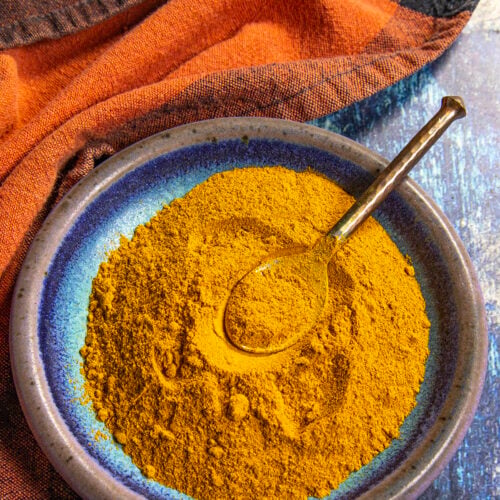 A blue ceramic plate filled with vibrant Ras el Hanout seasoning sits on a blue wooden surface, with a spoon resting in the golden spice and an orange cloth nearby.