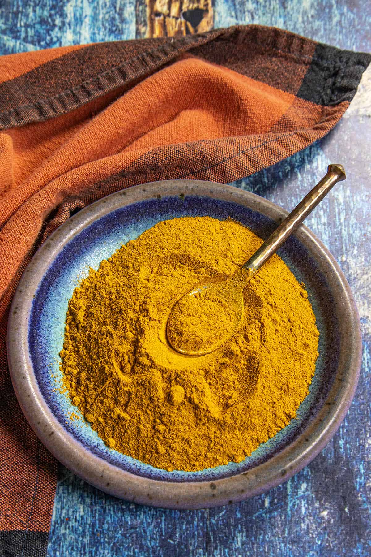 A blue ceramic plate filled with vibrant Ras el Hanout seasoning sits on a blue wooden surface, with a spoon resting in the golden spice and an orange cloth nearby.