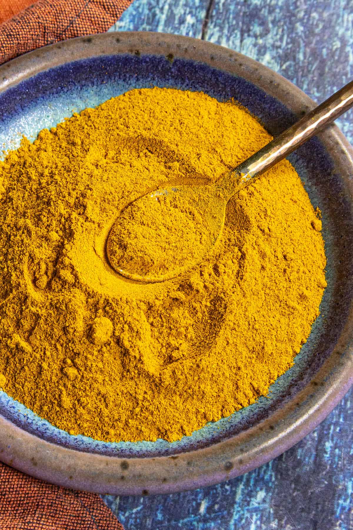 A ceramic plate filled with Homemade Ras el Hanout. A metal spoon rests in the powder, showing its bright orange-yellow color. The plate sits on a blue textured surface.