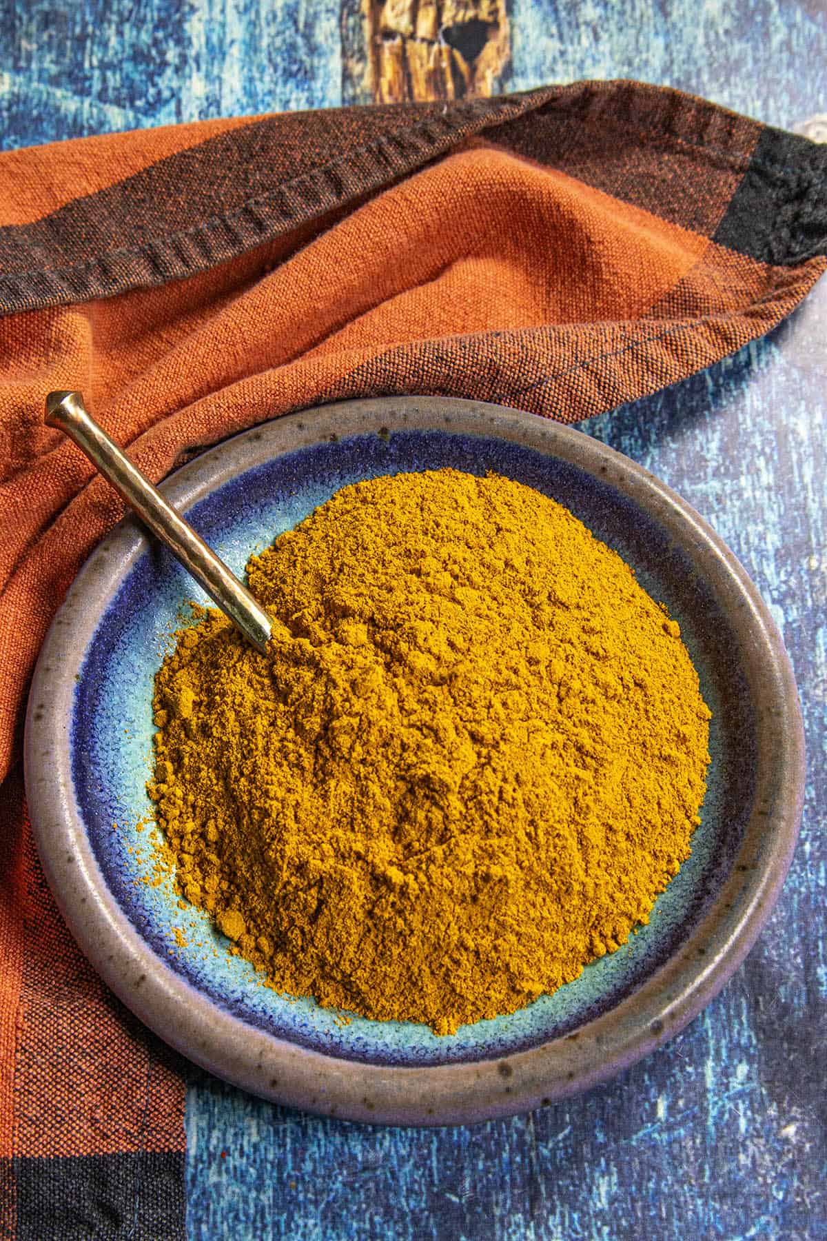 A blue ceramic plate with a pile of Homemade Ras el Hanout Seasoning curry powder and a gold spoon, placed on a blue surface near an orange and black checkered cloth.