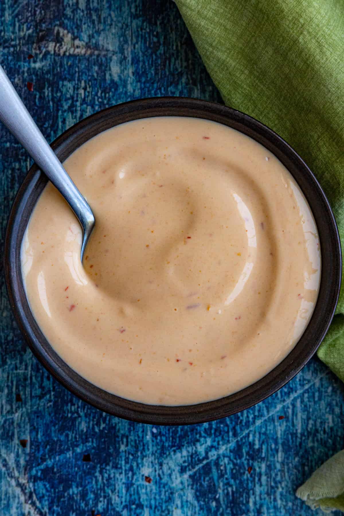 A bowl filled with creamy, light orange spicy mayo sits on a blue textured surface, with a spoon resting inside. A green cloth is partially visible to the side.