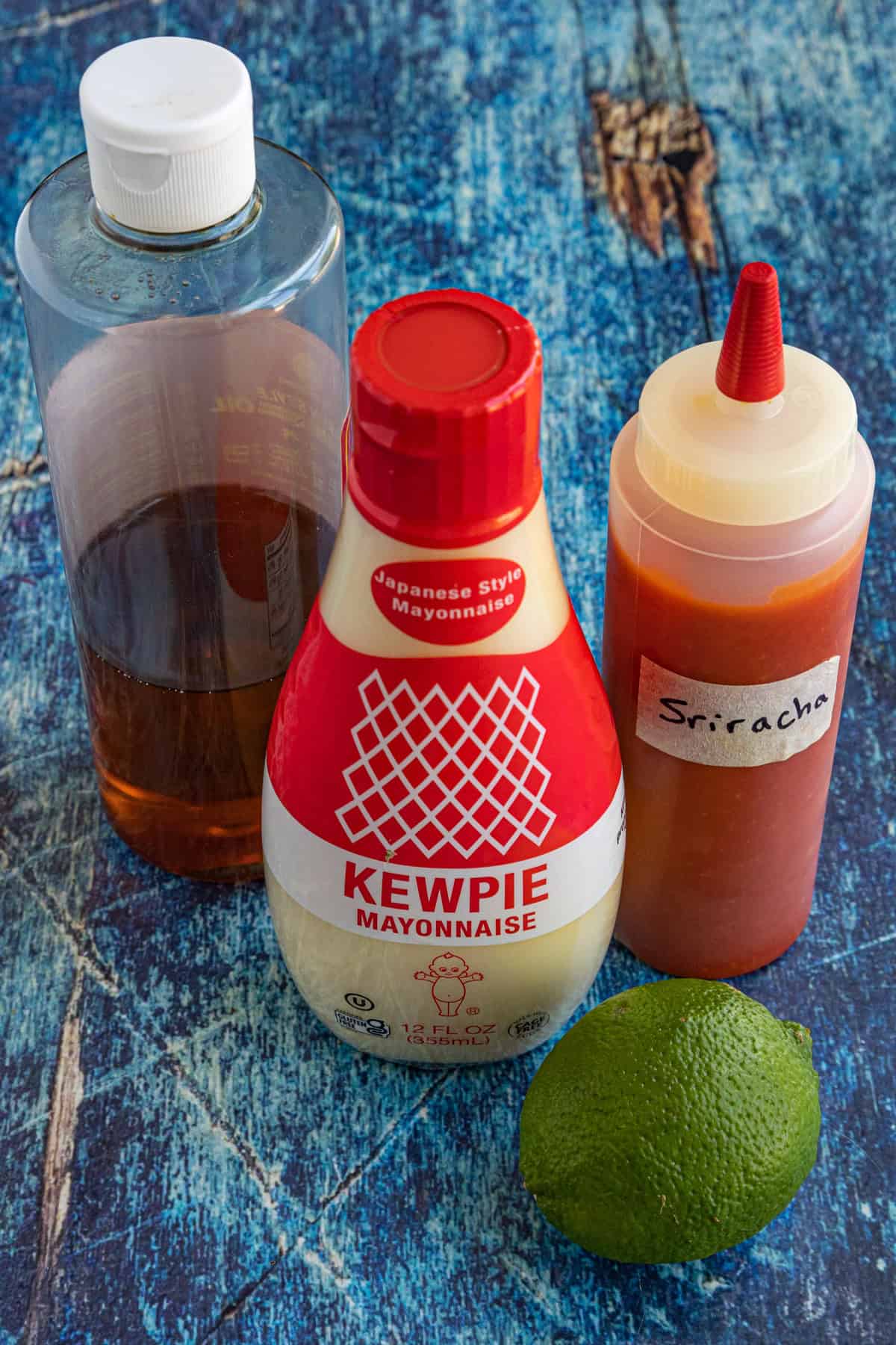 A bottle of sesame oil, Kewpie mayonnaise, a squeeze bottle of Sriracha for spicy mayo, and a fresh lime rest on a blue textured surface.
