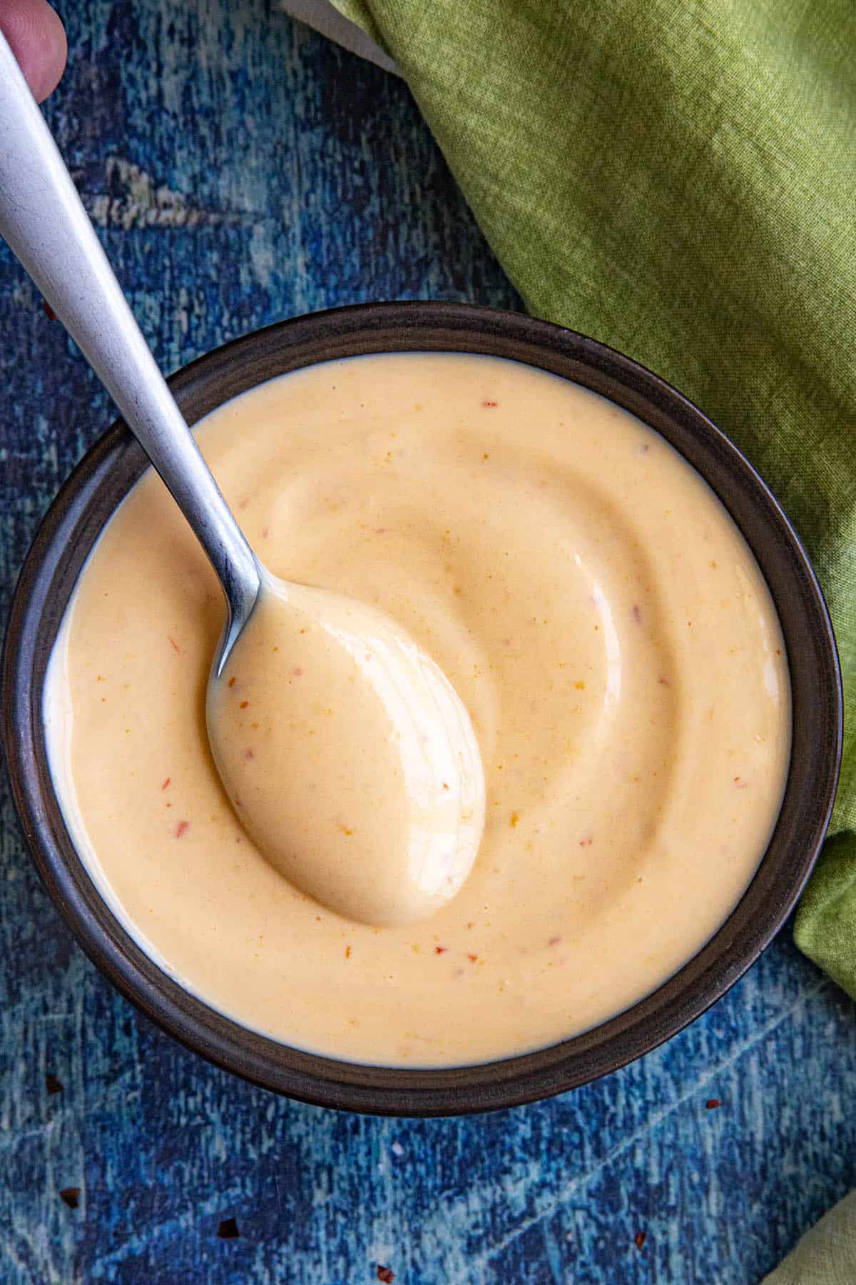 A spoon rests in a bowl filled with creamy, light orange spicy mayo on a blue textured surface, with a green cloth partially visible beside it.