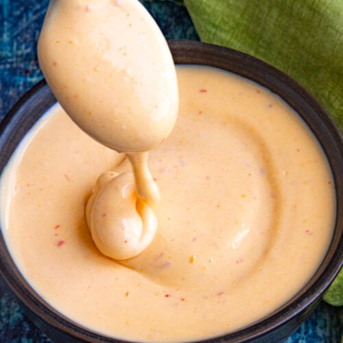 A spoon is dipped into a bowl of creamy, light orange spicy mayo with specks of seasoning, set on a blue textured surface near a green cloth.