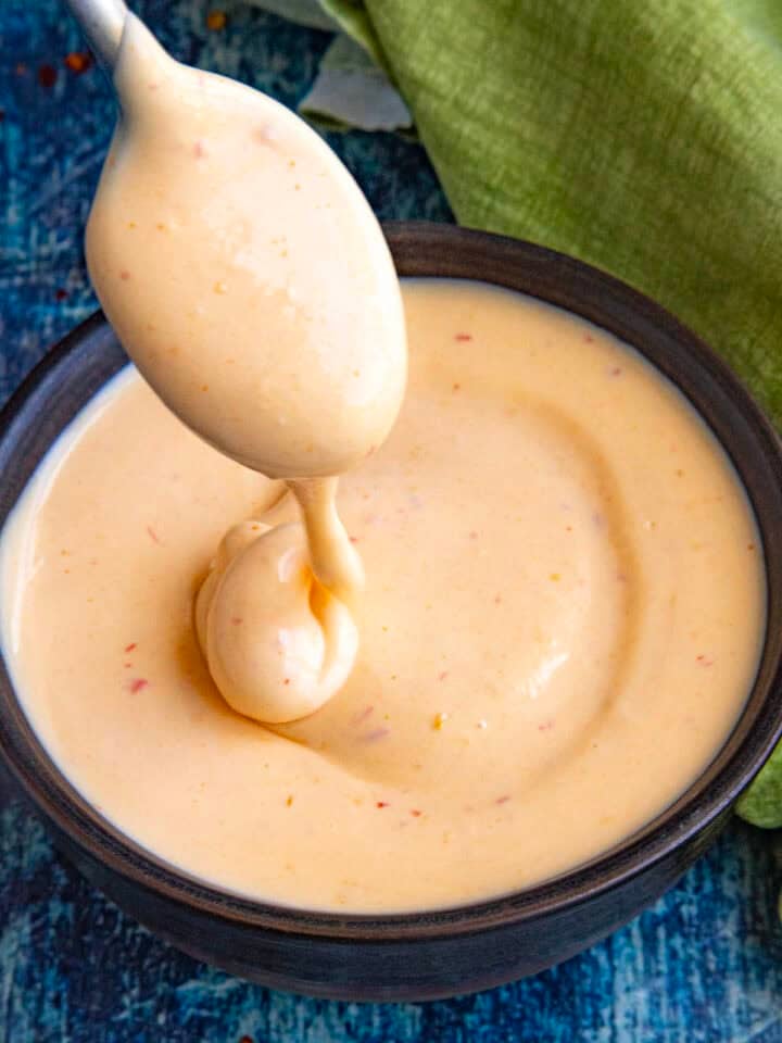 A spoon is dipped into a bowl of creamy, light orange spicy mayo with specks of seasoning, set on a blue textured surface near a green cloth.