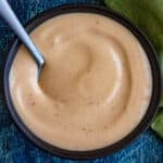 A bowl of creamy, light orange spicy mayo sauce with specks of seasoning, served with a spoon on a blue surface and a green cloth nearby.
