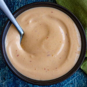 A bowl of creamy, light orange spicy mayo sauce with specks of seasoning, served with a spoon on a blue surface and a green cloth nearby.