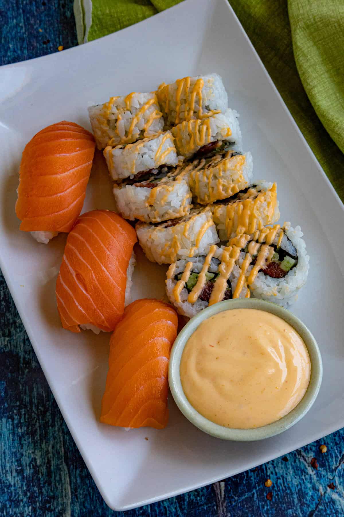 A white plate with four pieces of salmon nigiri, eight sushi rolls drizzled with spicy mayo, and a small bowl of creamy dipping sauce, set on a blue surface with a green napkin.