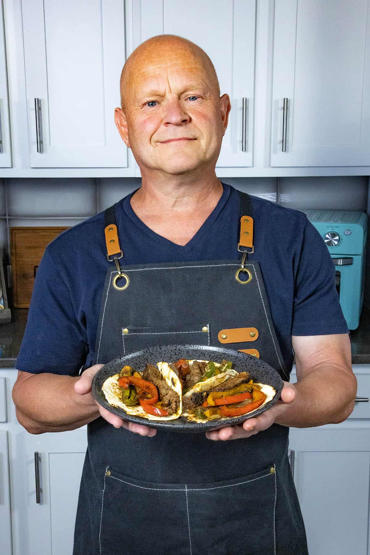 Mike Hultquist from Chili Pepper Madness wearing a dark apron stands in a kitchen, holding a plate with three steak fajitas filled with meat and vegetables. White cabinets and a teal kitchen appliance are visible in the background.