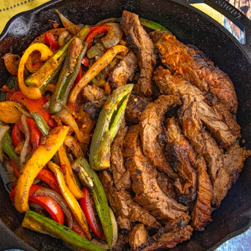 A cast iron skillet filled with cooked sliced beef, sautéed red, yellow, and green bell peppers, and onions—this Steak Fajitas Recipe is served on a blue wooden surface with a yellow cloth nearby.