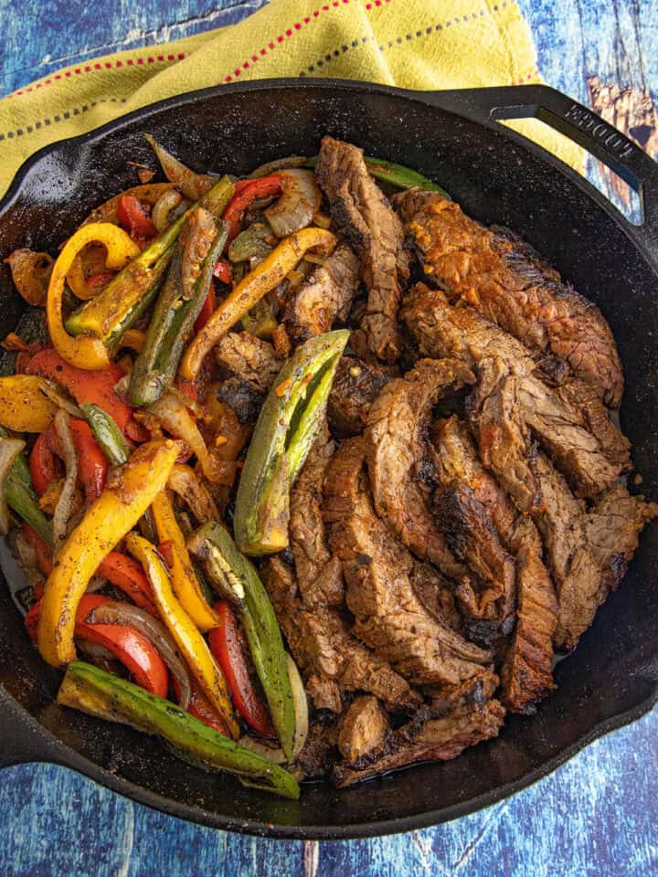 A cast iron skillet filled with cooked sliced beef, sautéed red, yellow, and green bell peppers, and onions—this Steak Fajitas Recipe is served on a blue wooden surface with a yellow cloth nearby.