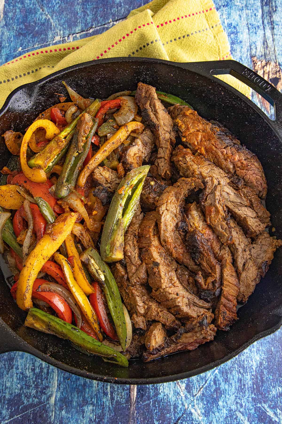 A cast iron skillet filled with cooked sliced beef, sautéed red, yellow, and green bell peppers, and onions—this Steak Fajitas Recipe is served on a blue wooden surface with a yellow cloth nearby.