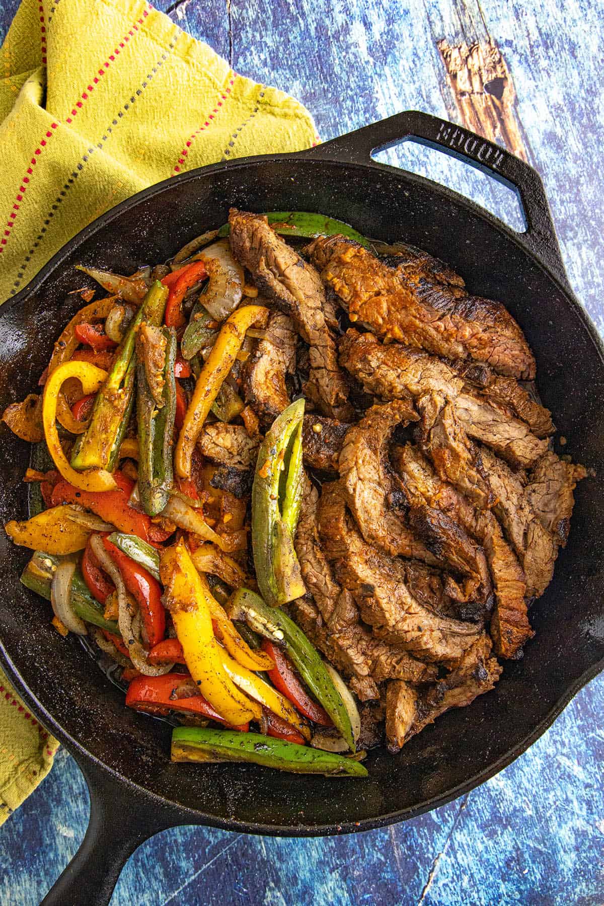 A cast iron skillet filled with sizzling Steak Fajitas&mdash;juicy sliced steak and saut&eacute;ed bell peppers and onions in vibrant colors&mdash;rests on a blue textured surface with a yellow patterned cloth nearby.