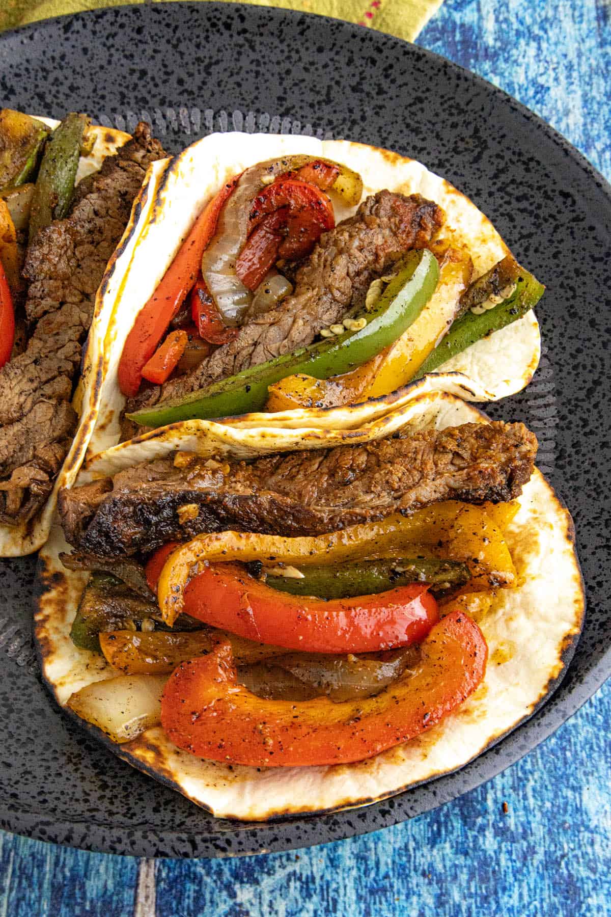 Three flour tortillas filled with spicy steak fajitas&mdash;grilled steak strips, red and green bell peppers, and saut&eacute;ed onions&mdash;are arranged on a dark plate set on a blue textured surface.
