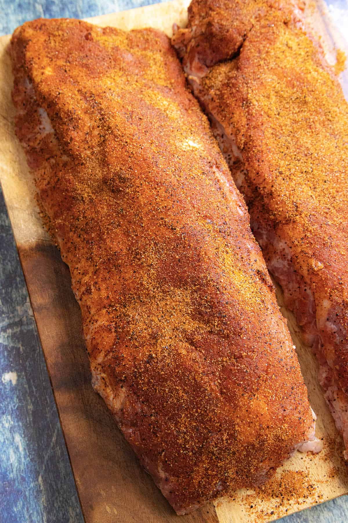 Two racks of uncooked pork ribs on a wooden cutting board, each coated with a generous layer of dry rub for ribs, set on a blue surface.