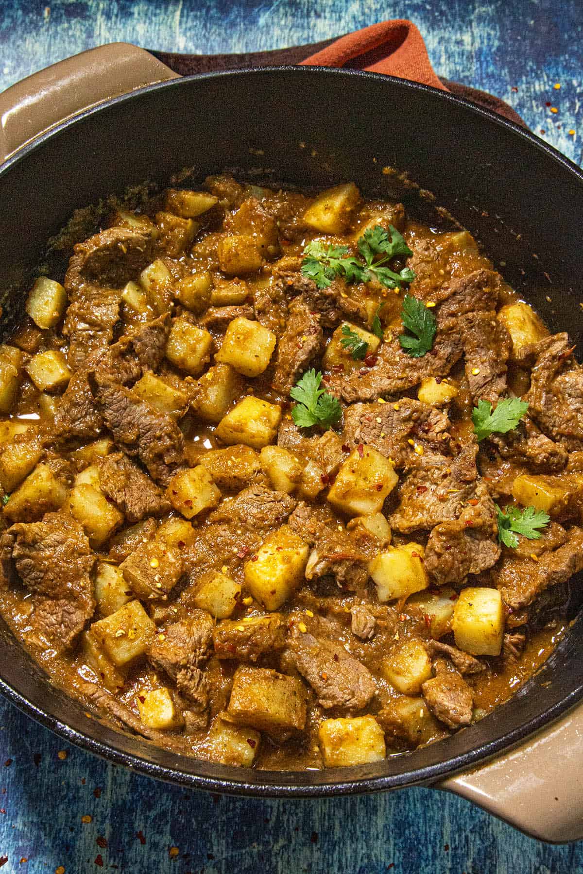 A pot filled with hearty Bistec Ranchero con Papas stew, garnished with fresh cilantro on top, set against a blue textured background.