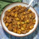 A bowl of Bistec Ranchero con Papas, featuring spicy beef stew with chunks of beef and potatoes in a rich, reddish-brown sauce, garnished with herbs, sits on a rustic blue wooden surface with a green napkin nearby.