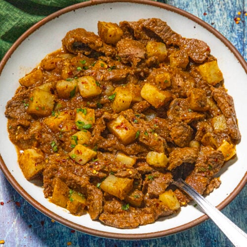 A bowl of beef caldereta, reminiscent of Bistec Ranchero con Papas, features chunks of beef, potatoes, and carrots in a rich, reddish-brown sauce, garnished with herbs. A spoon rests nearby on a green napkin atop a blue wooden surface.