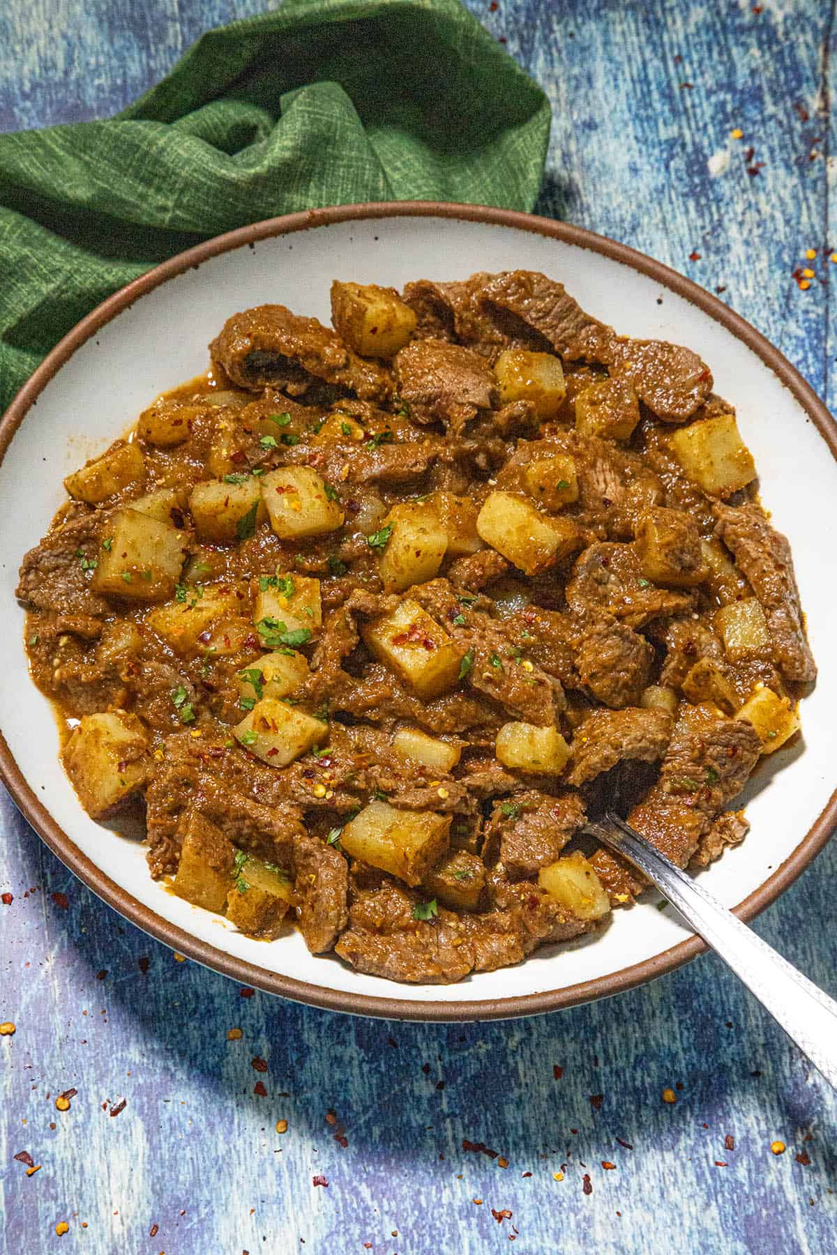 A bowl of beef caldereta, reminiscent of Bistec Ranchero con Papas, features chunks of beef, potatoes, and carrots in a rich, reddish-brown sauce, garnished with herbs. A spoon rests nearby on a green napkin atop a blue wooden surface.