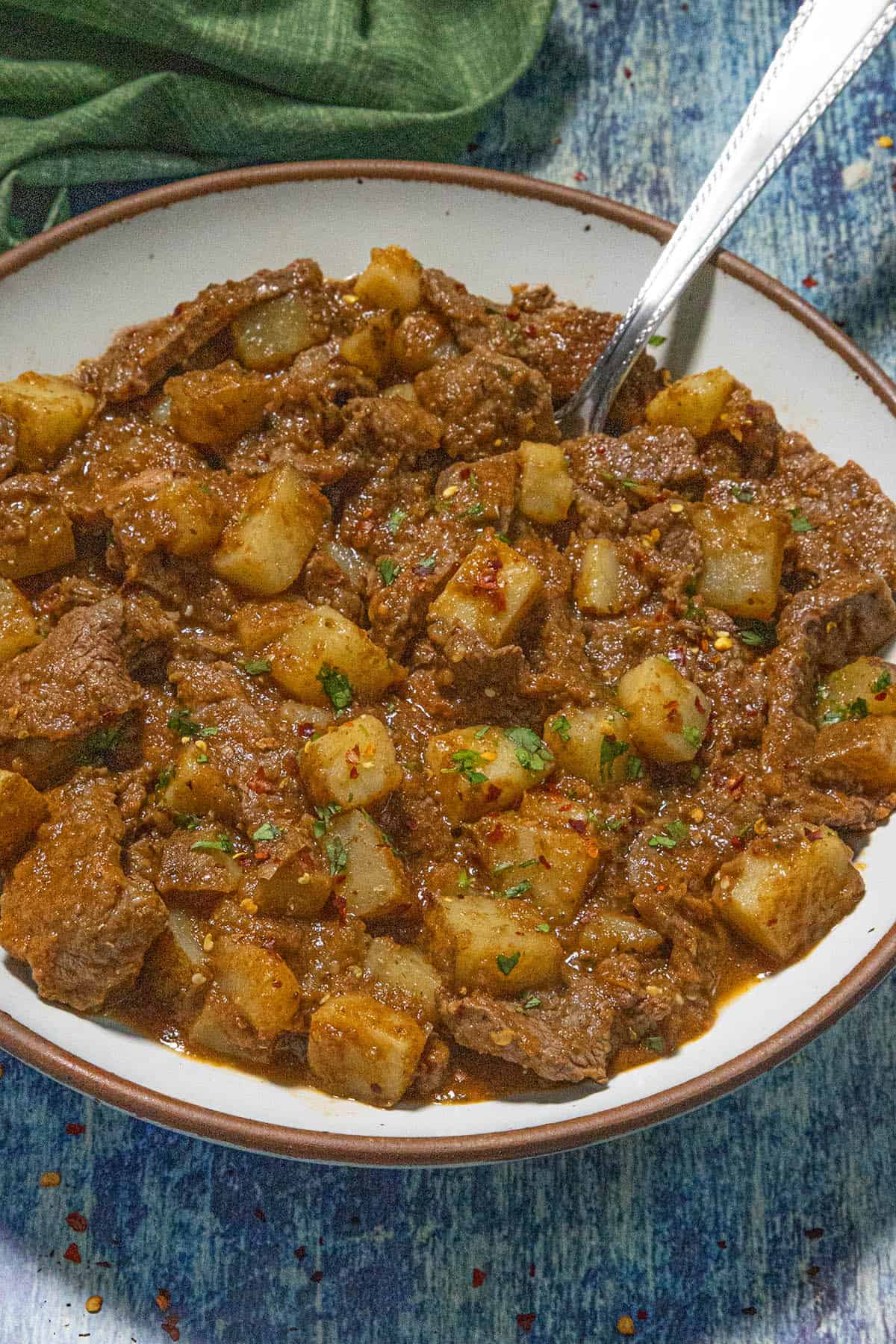 A bowl of Bistec Ranchero with potatoes, garnished with herbs and red pepper flakes, sits on a blue textured surface with a spoon in the bowl.