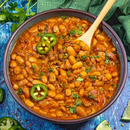 A bowl of spicy Borracho Beans (Frijoles Borrachos) topped with sliced jalapeños and fresh cilantro, with a wooden spoon inside. Lime wedges, jalapeño slices, and cilantro are scattered on a blue wooden surface around the bowl.