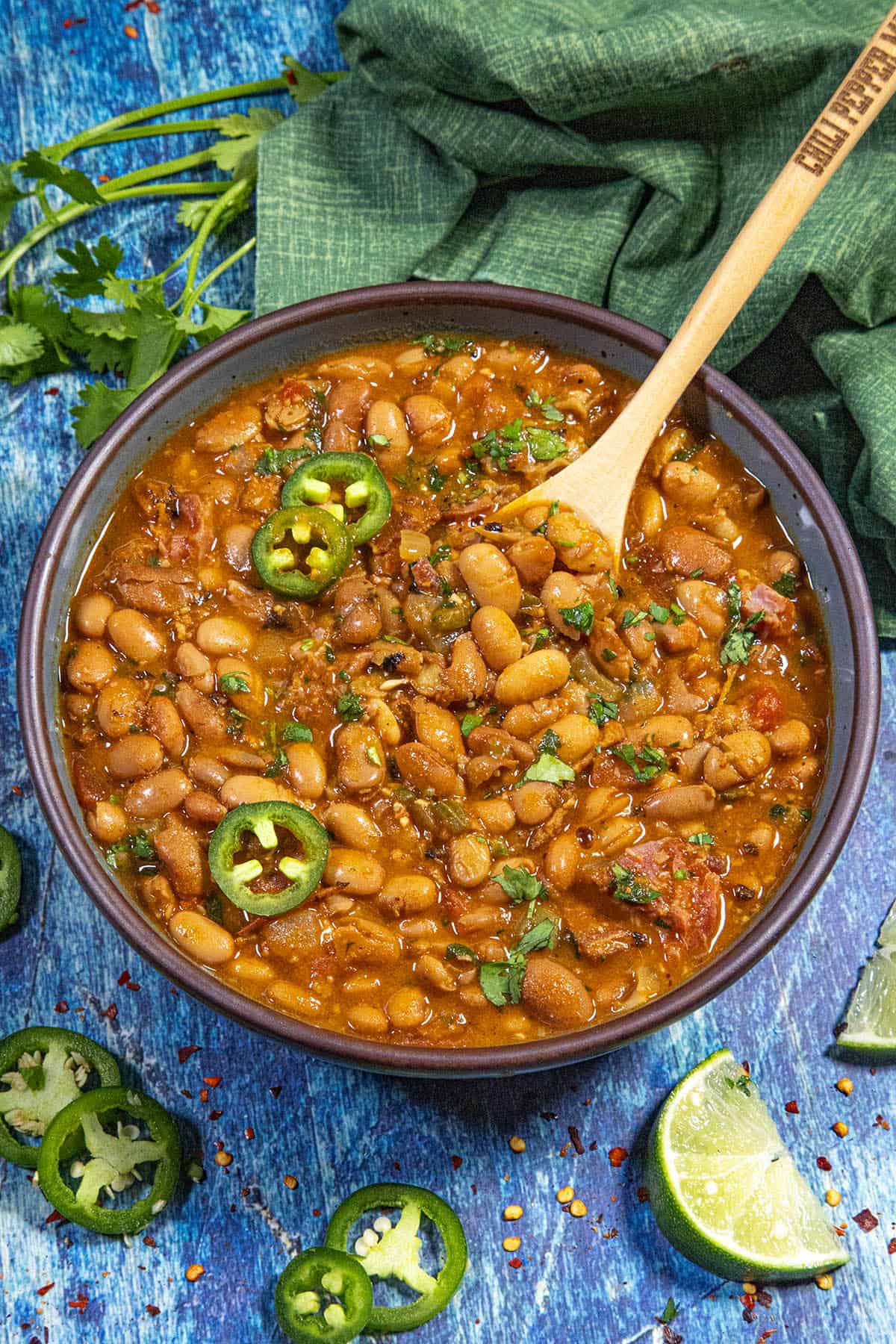 A bowl of spicy Borracho Beans (Frijoles Borrachos) topped with sliced jalapeños and fresh cilantro, with a wooden spoon inside. Lime wedges, jalapeño slices, and cilantro are scattered on a blue wooden surface around the bowl.
