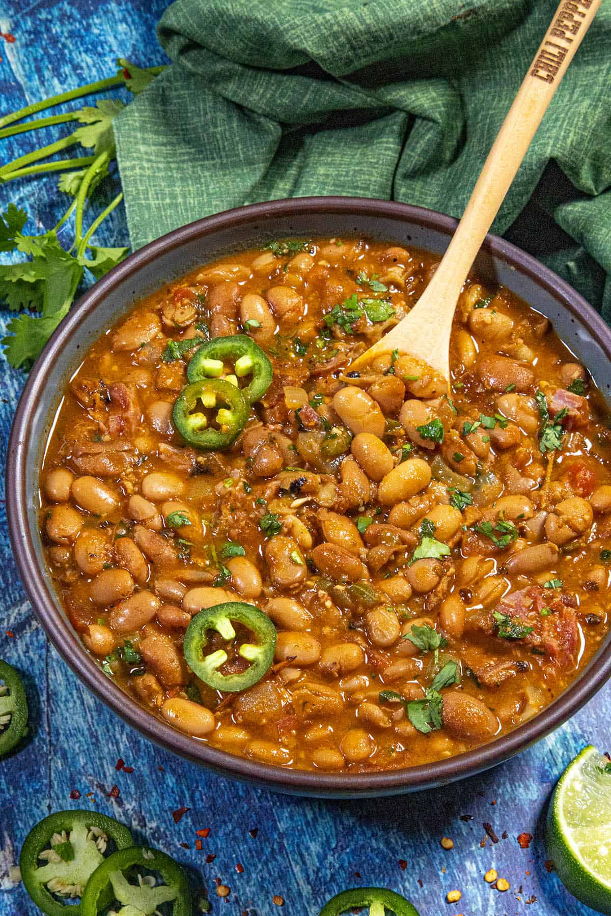 A bowl of spicy Borracho Beans (Frijoles Borrachos) garnished with sliced jalapeños and chopped cilantro, with a wooden spoon resting in the bowl. Fresh cilantro and jalapeños are scattered nearby on a blue surface.