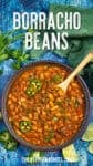 A bowl of Borracho Beans garnished with cilantro and sliced jalapeños sits ready with a wooden spoon. Fresh cilantro, sliced jalapeños, lime wedges, and a green napkin are nearby on a blue textured surface.