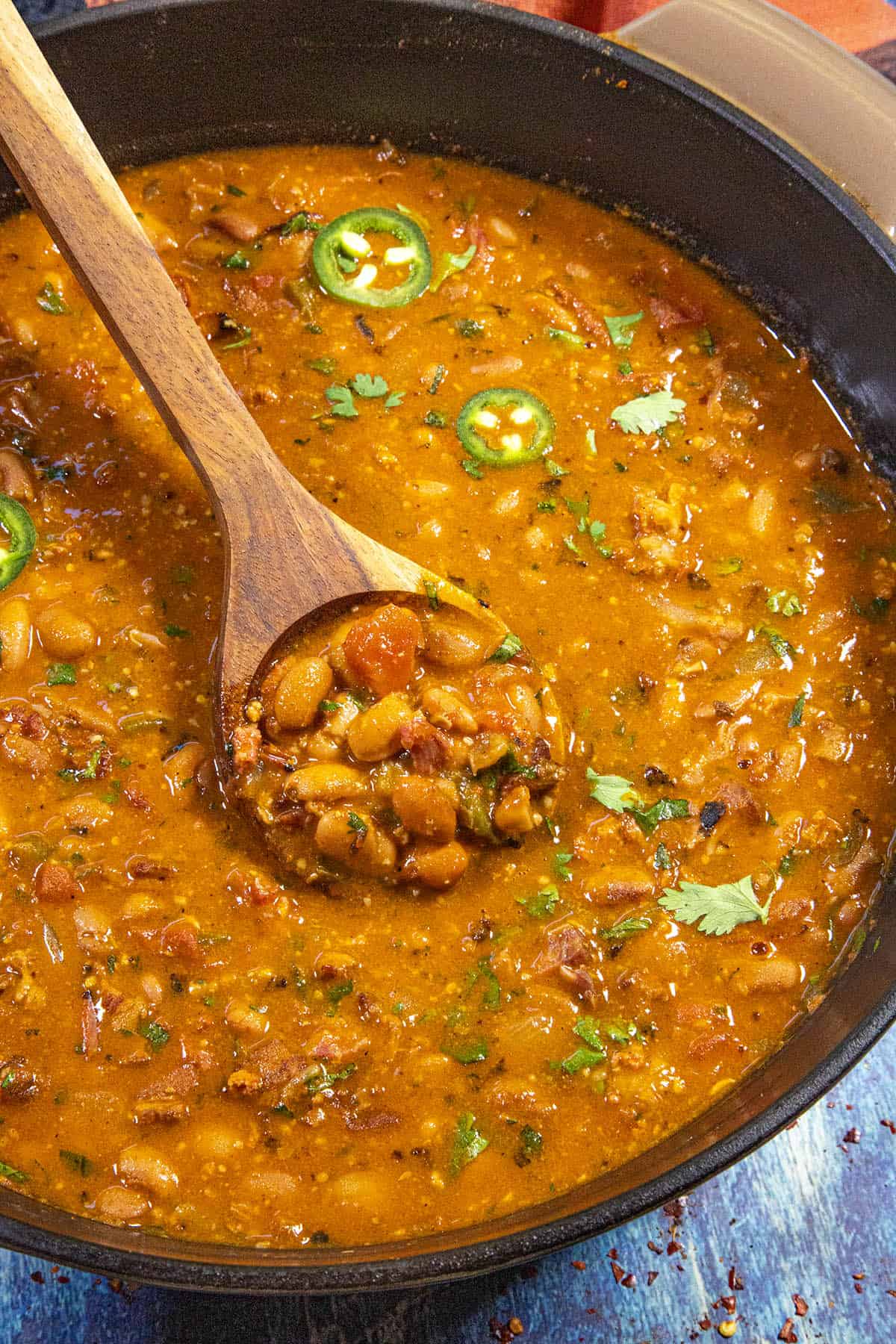 A pot of spicy bean chili inspired by Frijoles Borrachos (Mexican Borracho Beans), topped with sliced jalapeños and cilantro, with a wooden spoon lifting a portion of the thick, hearty mixture.