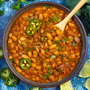 A bowl of Frijoles Borrachos (Mexican Borracho Beans) garnished with sliced jalapeños and chopped cilantro, with a wooden spoon. Lime wedges and jalapeño slices are scattered on the blue wooden surface around the bowl.