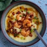 A bowl of creamy grits topped with seasoned shrimp, crumbled bacon, chopped green onions, and a drizzle of sauce&mdash;a Classic Shrimp and Grits Recipe&mdash;served with a spoon resting inside on a blue textured surface.