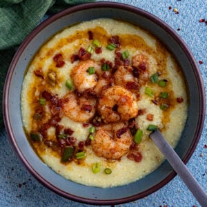A bowl of creamy grits topped with seasoned shrimp, crumbled bacon, chopped green onions, and a drizzle of sauce&mdash;a Classic Shrimp and Grits Recipe&mdash;served with a spoon resting inside on a blue textured surface.