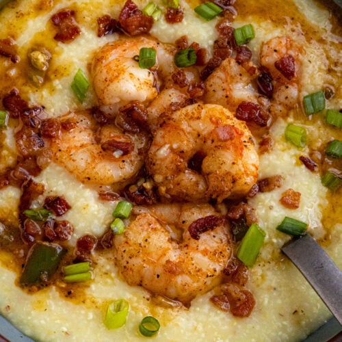 A bowl of Classic Shrimp and Grits topped with seasoned shrimp, crispy bacon pieces, chopped green onions, and a drizzle of sauce, with a spoon resting inside.