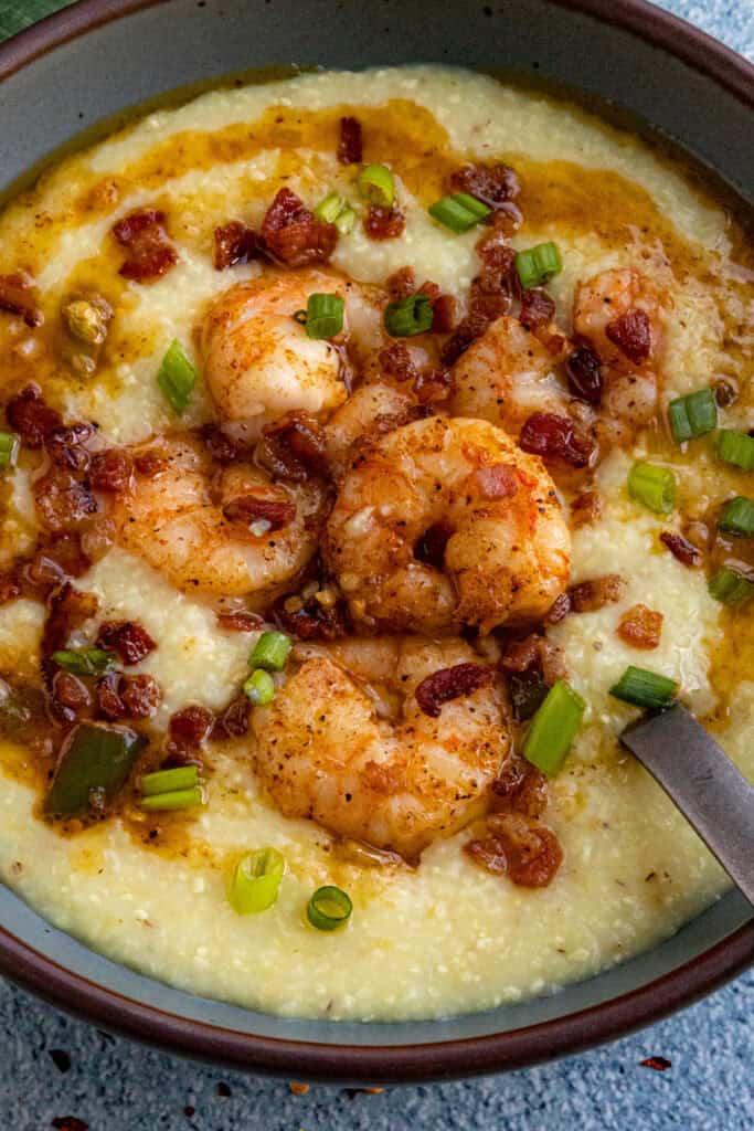 A bowl of Classic Shrimp and Grits topped with seasoned shrimp, crispy bacon pieces, chopped green onions, and a drizzle of sauce, with a spoon resting inside.