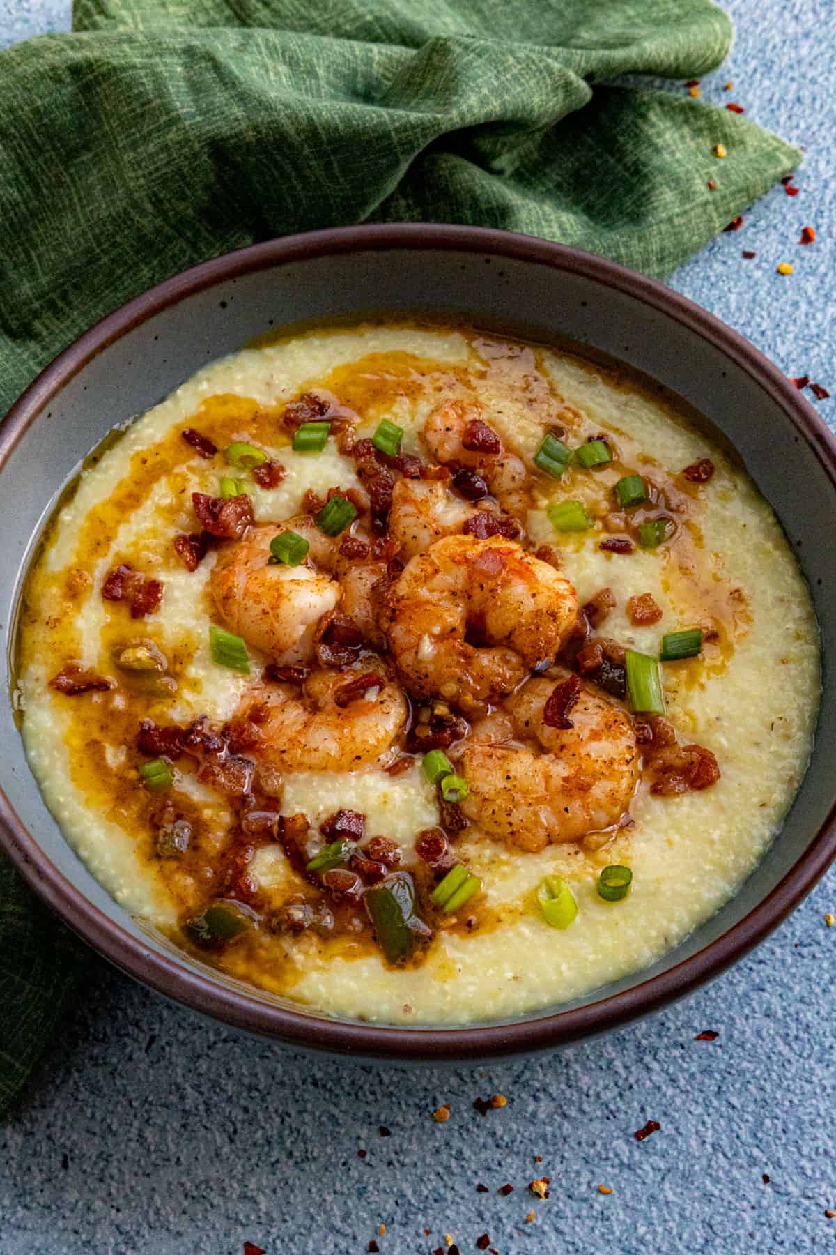 A bowl of Classic Southern Shrimp and Grits topped with shrimp, crumbled bacon, chopped green onions, and a drizzle of sauce sits on a blue surface with a green cloth nearby.