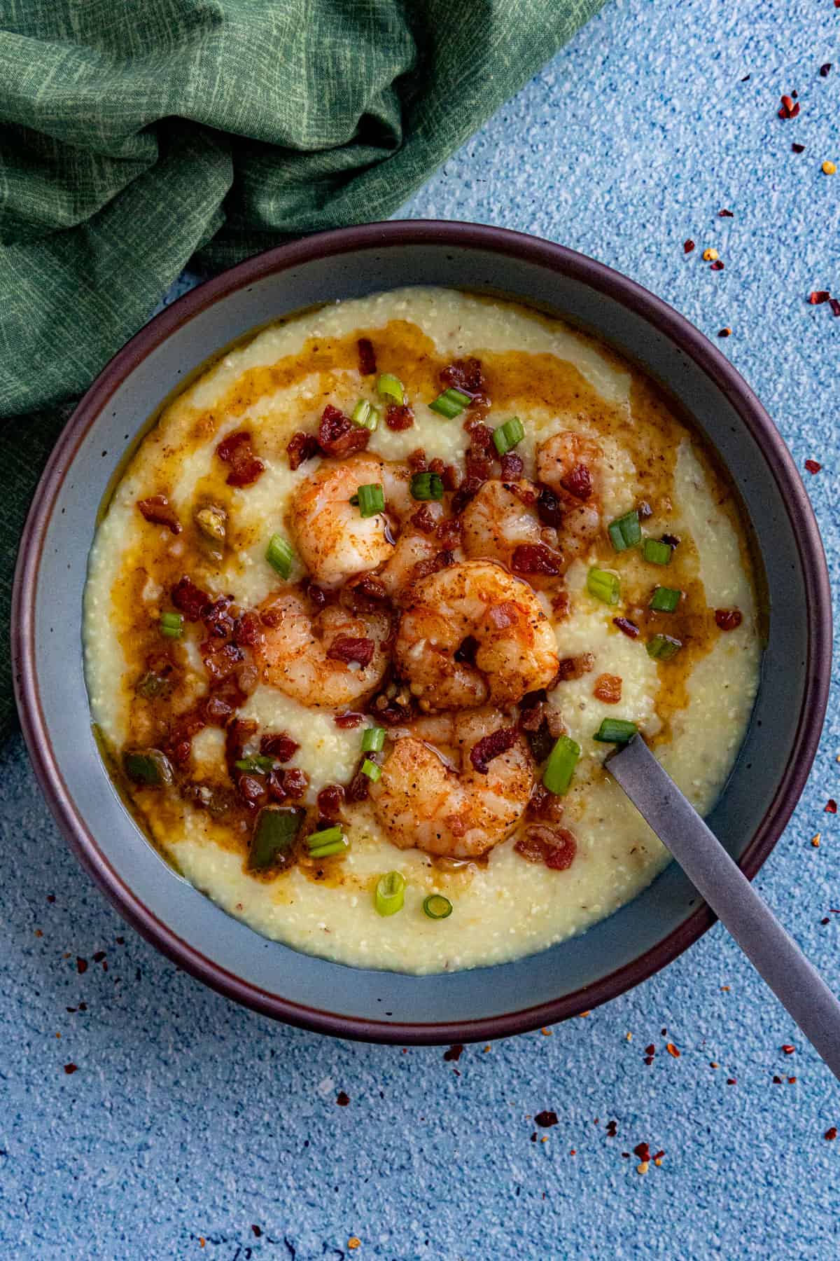 A bowl of creamy Shrimp and Grits topped with crispy bacon bits, sliced green onions, and a drizzle of sauce, with a spoon resting in the bowl on a blue textured surface next to a green cloth.