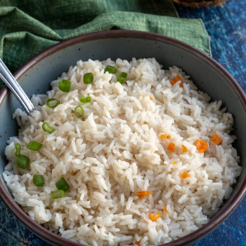 A bowl of Creamy Coconut Rice garnished with chopped green onions and small habanero pieces, with a spoon resting inside. A green cloth and half a coconut are in the background on a blue surface.