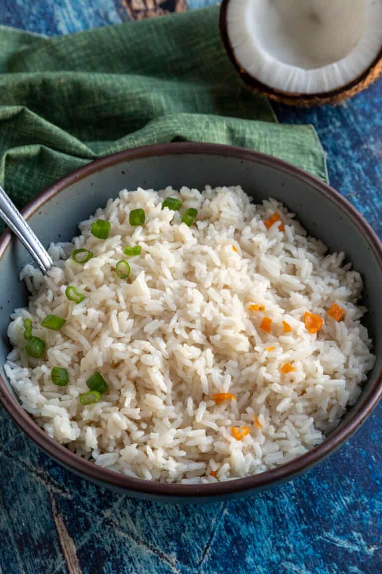 A bowl of Creamy Coconut Rice garnished with chopped green onions and small habanero pieces, with a spoon resting inside. A green cloth and half a coconut are in the background on a blue surface.