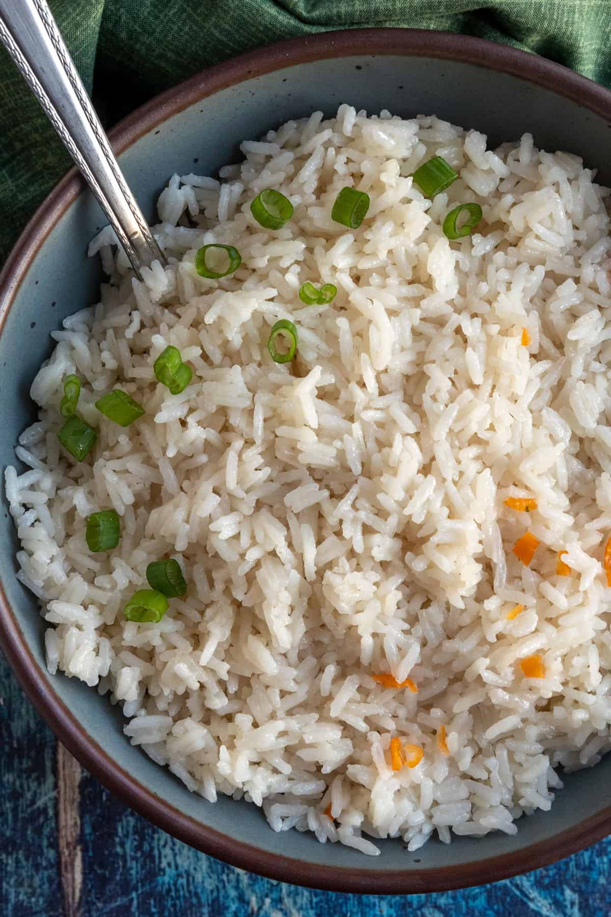 A bowl of cooked coconut rice garnished with chopped green onions and a few small orange vegetable pieces, with a fork resting in the bowl.